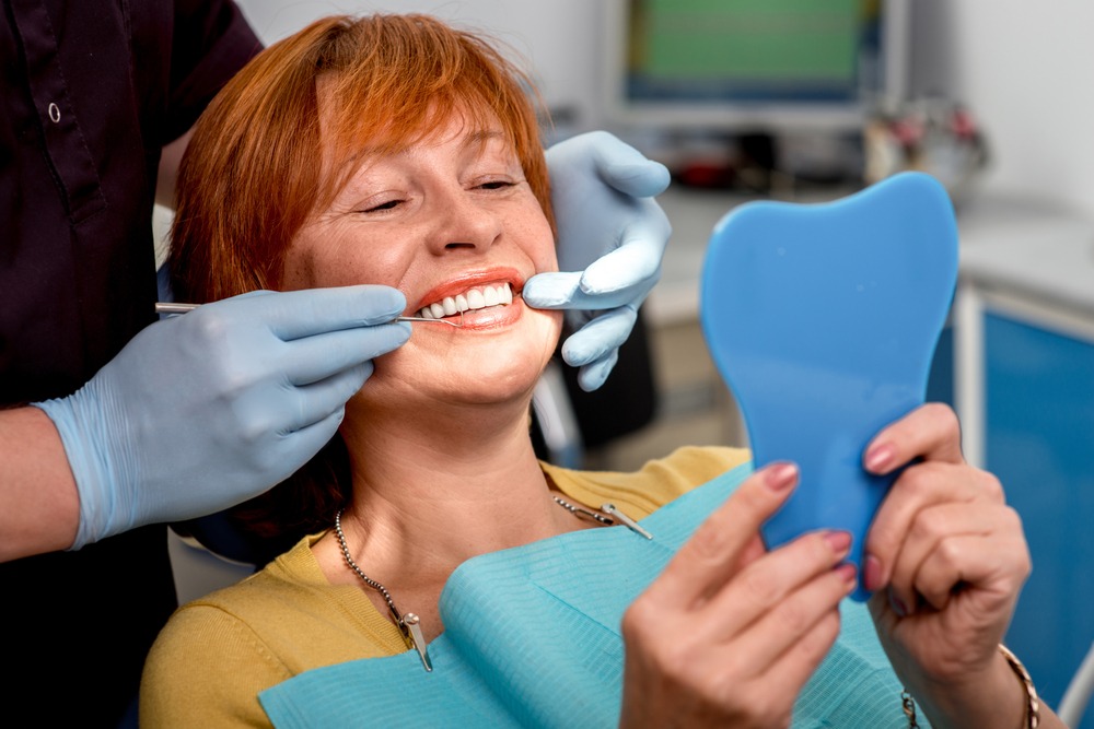 Older woman in a dental chair checking out her new dentures in a mirror.